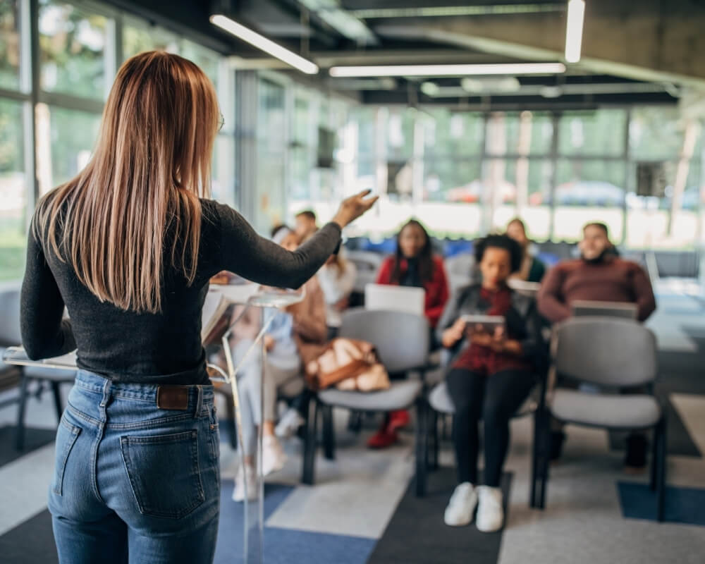 A woman speaking and a group of people sitting and listening to her on chairs.