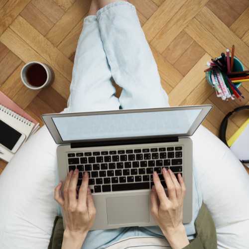 A person sitting with a laptop in their lap, a coffee cup placed on the left side of them and a pen pot on the right side.